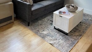 Una sala de estar acogedora con un sofá gris oscuro, una mesita auxiliar blanca y un ventanal con persianas.