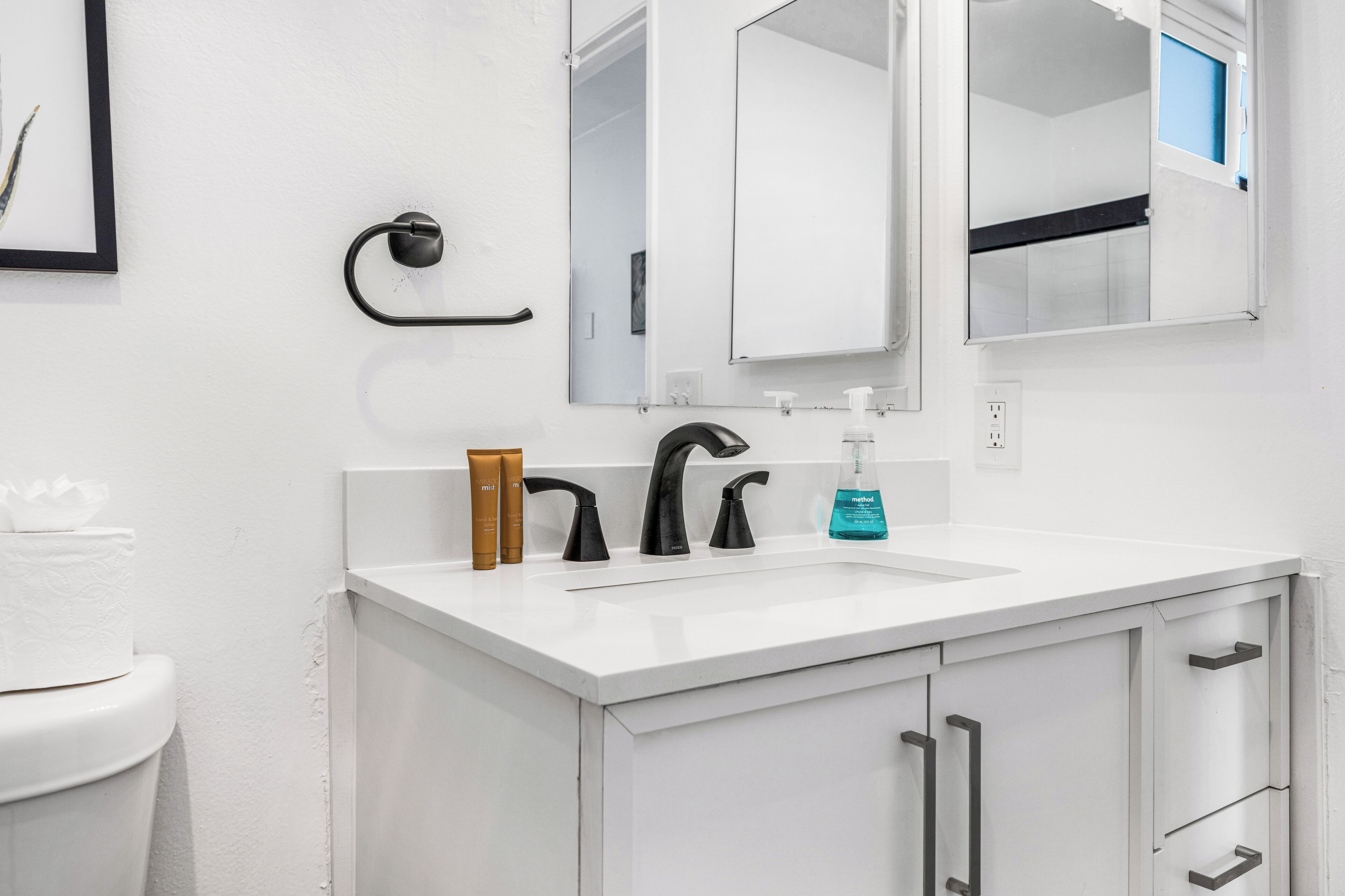 Un baño moderno con un lavamanos blanco, grifos negros y un espejo encima. Hay artículos de aseo en la encimera y una toalla en el suelo.