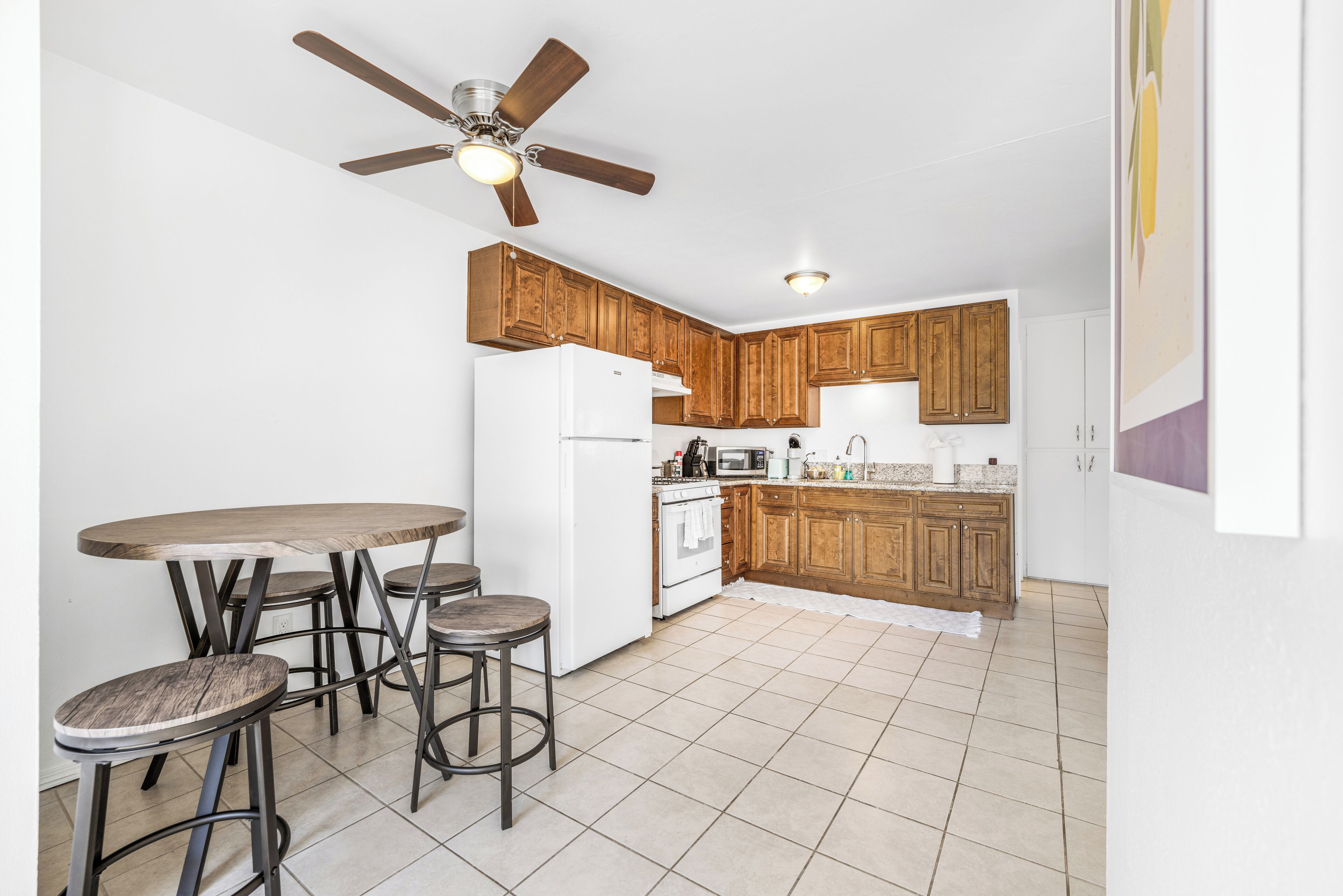 Une cuisine avec des meubles de rangement en bois, un réfrigérateur blanc, une cuisinière et un ventilateur de plafond. Il y a quatre tabourets autour d’une table.