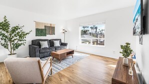 A modern living room with a grey sofa, wooden coffee table, and a large window offering a view of palm trees and buildings. - NEW 1br Ocean Beach Apartment w/ Parking - Short Walk to the Beach (San Diego)