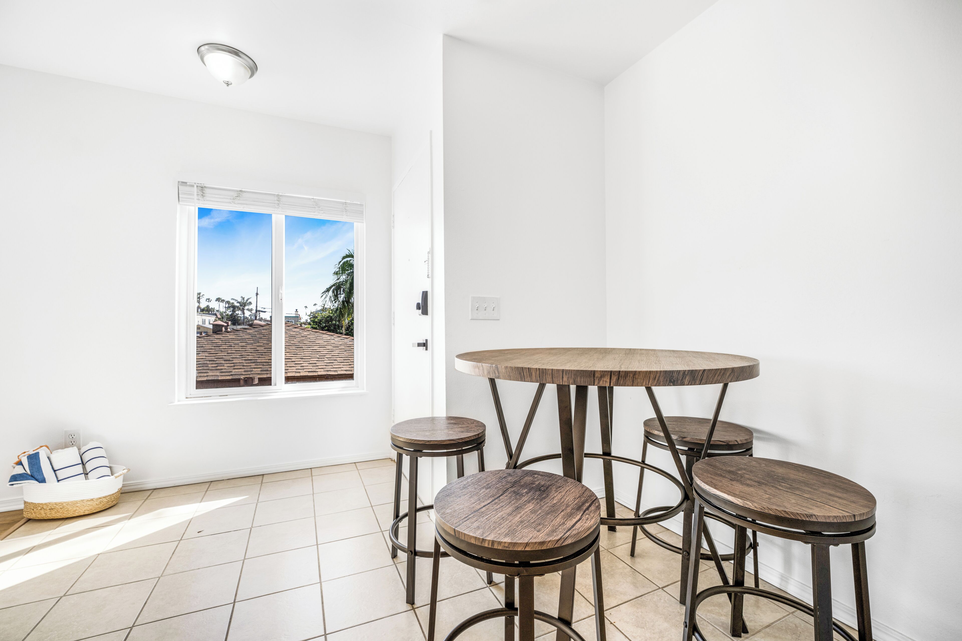 A room with a round wooden table and four stools, a white door, and a window with a view of a building and trees.