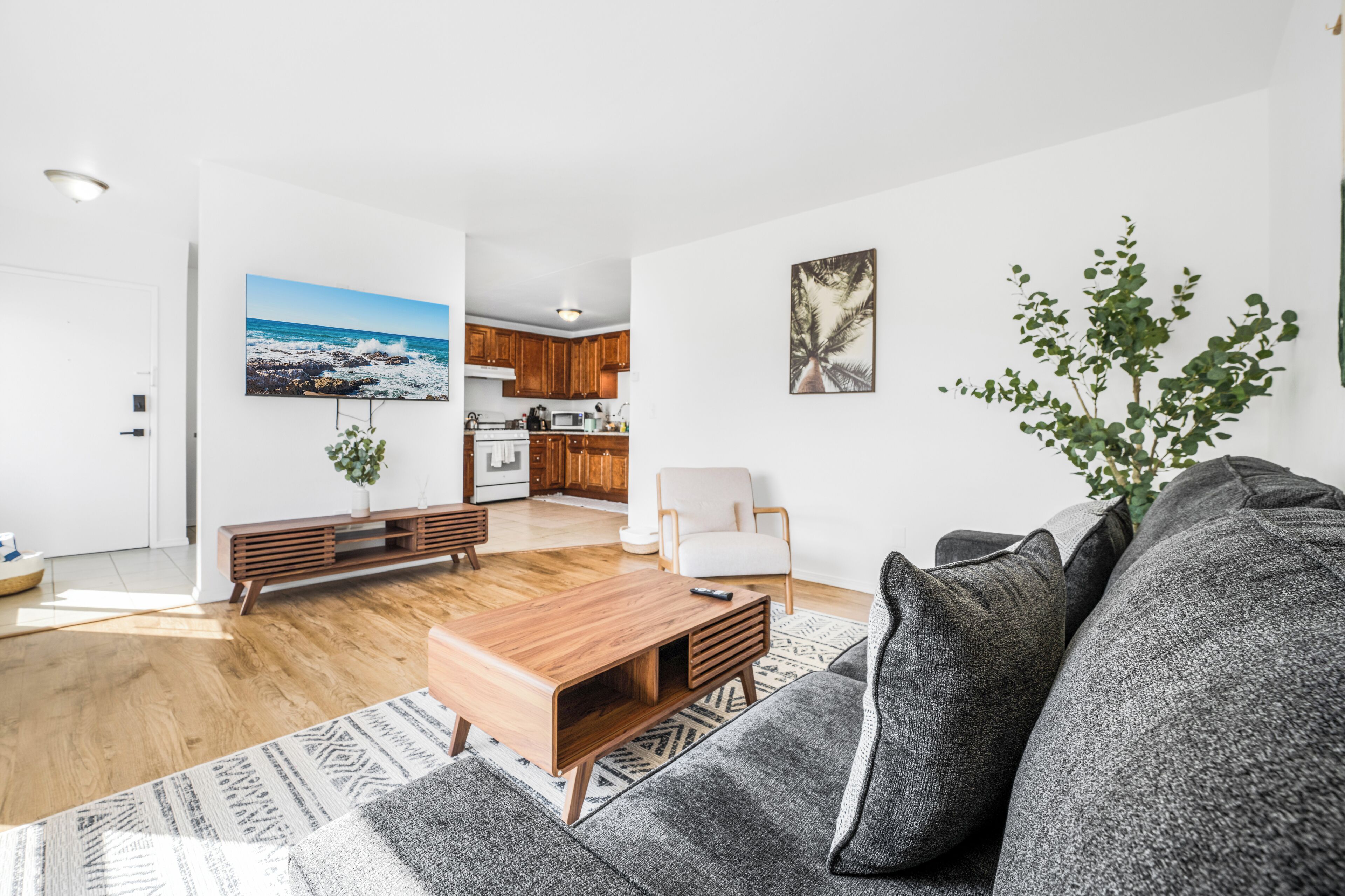 A modern living room with a grey sofa, wooden coffee table, and a white armchair. A kitchen with wooden cabinets and a dining area is visible in the background.