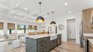 A modern kitchen with a central island, pendant lighting, and a view of the living area. - Exceptional New Build - West End - Pool - Golf Cart (Panama City Beach)