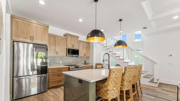 A modern kitchen with wooden cabinets, stainless steel appliances, a central island with bar stools, and a staircase leading to an upper level. - Exceptional New Build - West End - Pool - Golf Cart (Panama City Beach)