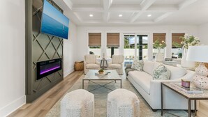 A modern living room with a flat-screen TV mounted on a wall, a sofa, two armchairs, a coffee table, and a large window with blinds. - Exceptional New Build - West End - Pool - Golf Cart (Panama City Beach)
