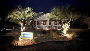 A house with a sign reading "OKIDDUK" surrounded by palm trees and a wooden fence.