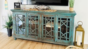 A teal-colored wooden console table with glass doors, a wicker basket, and a small potted plant.