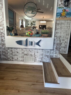 A living room with a large fish decoration, a ceiling fan, and a beach-themed painting. - Cozy 4 Bedroom beach bungalow with private HEATED pool & golf cart. (Oak Island)
