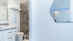 Une salle de bain moderne avec un mur de douche en marbre, un lavabo blanc doté d’un plan de toilette en marbre et un grand miroir avec une bande lumineuse bleue.