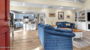 A spacious living area with a blue sofa, a dining table, and a kitchen in the background. - 5 mins to beach, golf cart & fenced in yard. Pet-friendly! (Oak Island)