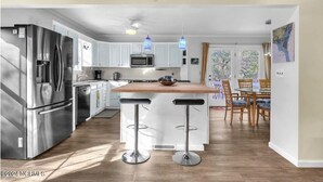A modern kitchen with stainless steel appliances, a central island with bar stools, and a dining area with wooden furniture. - 5 mins to beach, golf cart & fenced in yard. Pet-friendly! (Oak Island)