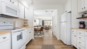 Eine moderne Küche mit weißen Schränken, einer Mikrowelle, einem Herd und einem Kühlschrank. Im Hintergrund ist eine Essecke mit Tisch und Stühlen zu sehen.