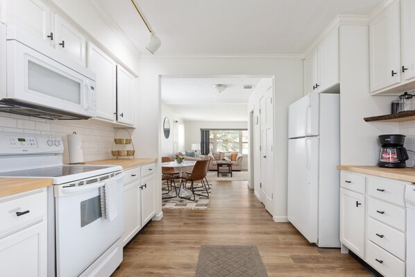 Une cuisine moderne avec des meubles blancs, un four à micro-ondes, une cuisinière et un réfrigérateur. On aperçoit à l’arrière un coin repas avec une table et des chaises.