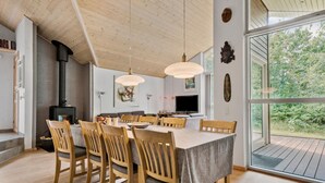 Uma sala de jantar com uma mesa grande, cadeiras de madeira, luminárias pendentes, lareira e uma grande janela com vista para o verde.