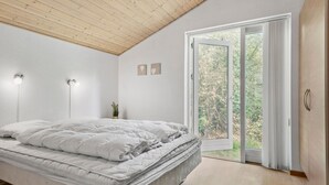 Quarto com cama, duas luzes fixadas na parede, um vaso de planta e uma porta de vidro deslizante que dá para uma varanda com vista para árvores.