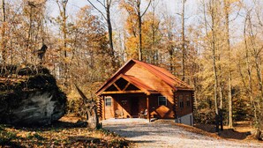Une cabane en bois dotée d’un porche, entourée d’arbres et bordée d’un grand rocher.