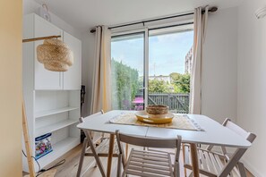 A dining area with a white table, wooden chairs, a woven light fixture, and a view of greenery and buildings outside. - Appartement Chanzy - Welkeys (Arcachon)