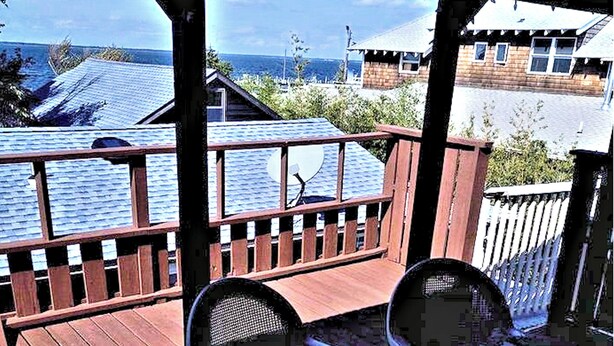 A wooden deck with two chairs, a view of the sea, and a building with a sloped roof in the background. - Ocean Beach, Fire Island,NY 4 bed,2 Bath, Sleeps 9, Full Kitchen, Deck, Bicycles (Ocean Beach)