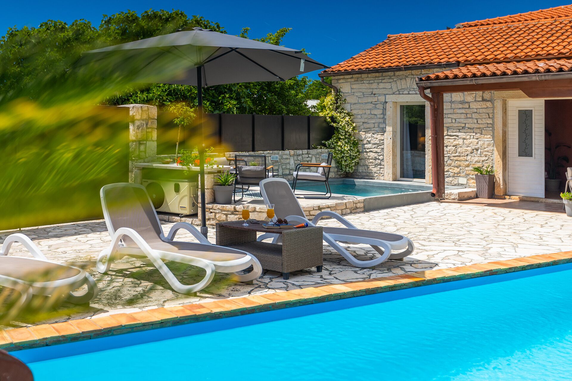 Area piscina con poltrone relax, un tavolo e un edificio in pietra con tetto in tegole.