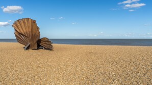 A large metallic sculpture of a shell on a pebbled beach with a clear sky.