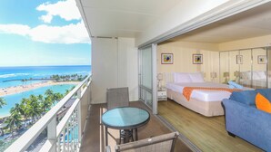A balcony with a glass table and chairs, a bed with a blue and orange pillow, and a view of the beach and ocean.