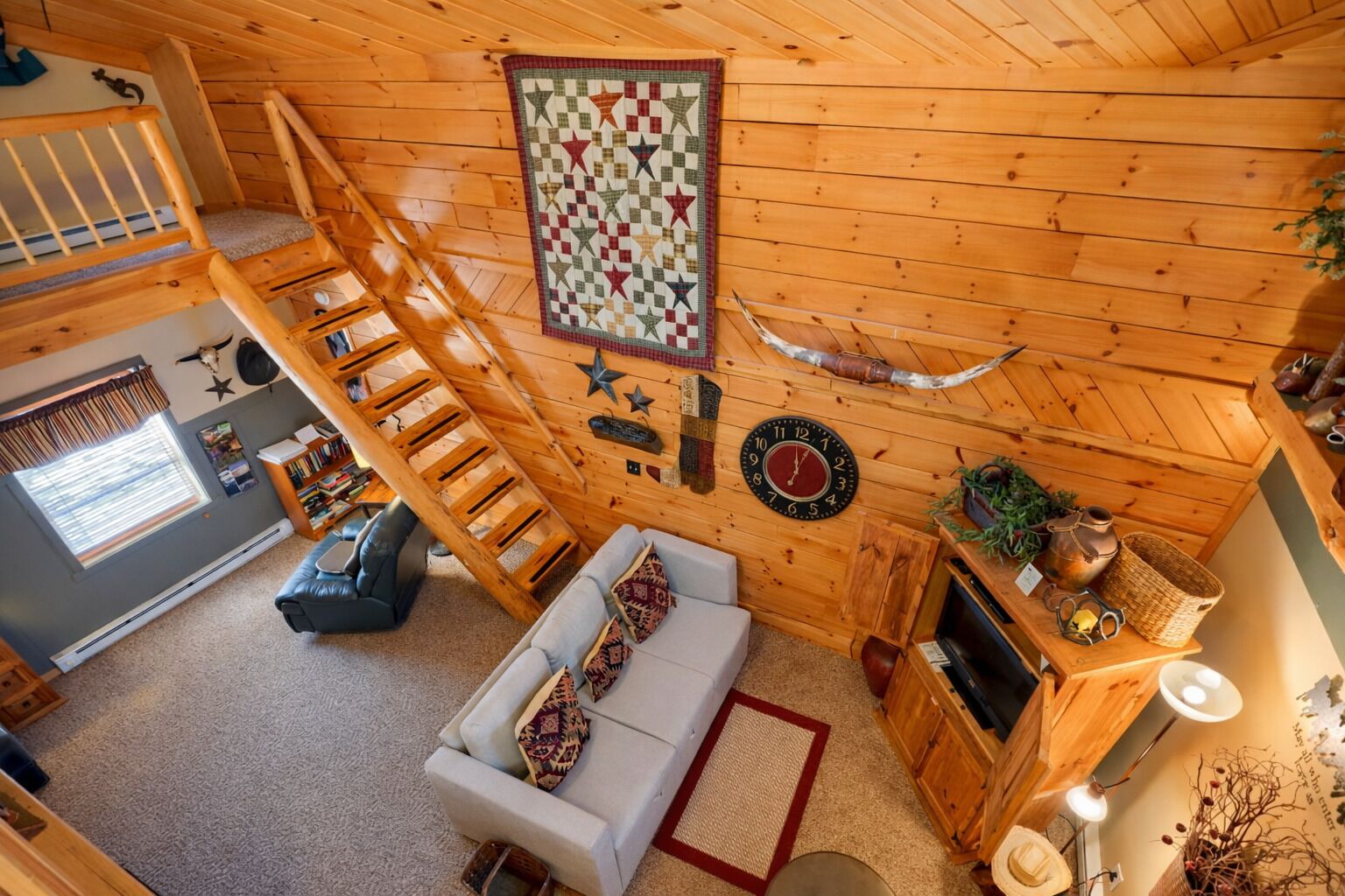 A cozy cabin interior with a loft bedroom, a living area with a sofa and a fireplace, and a wooden staircase.