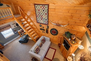 A cozy cabin interior with a loft bedroom, a living area with a sofa and a fireplace, and a wooden staircase.