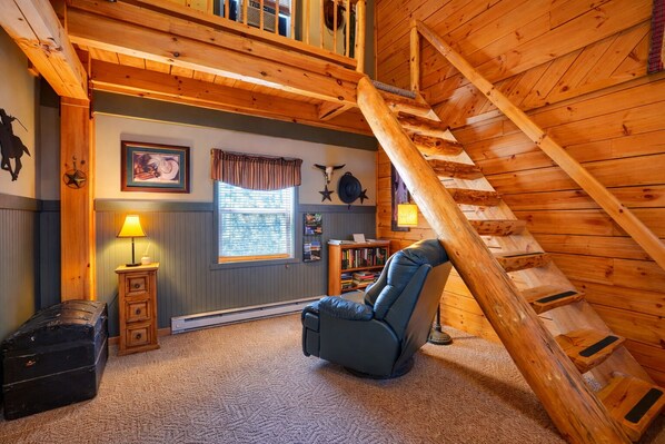 A cozy room with a sloped ceiling, a wooden staircase, a comfortable chair, a small table with a lamp, and a bookshelf.