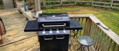A barbecue grill on a wooden deck with a metal table and a chair nearby.