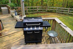 A barbecue grill on a wooden deck with a metal table and a chair nearby. - Carp's Cabin: 2 Bedroom Farmhouse Bungalow (Logan)
