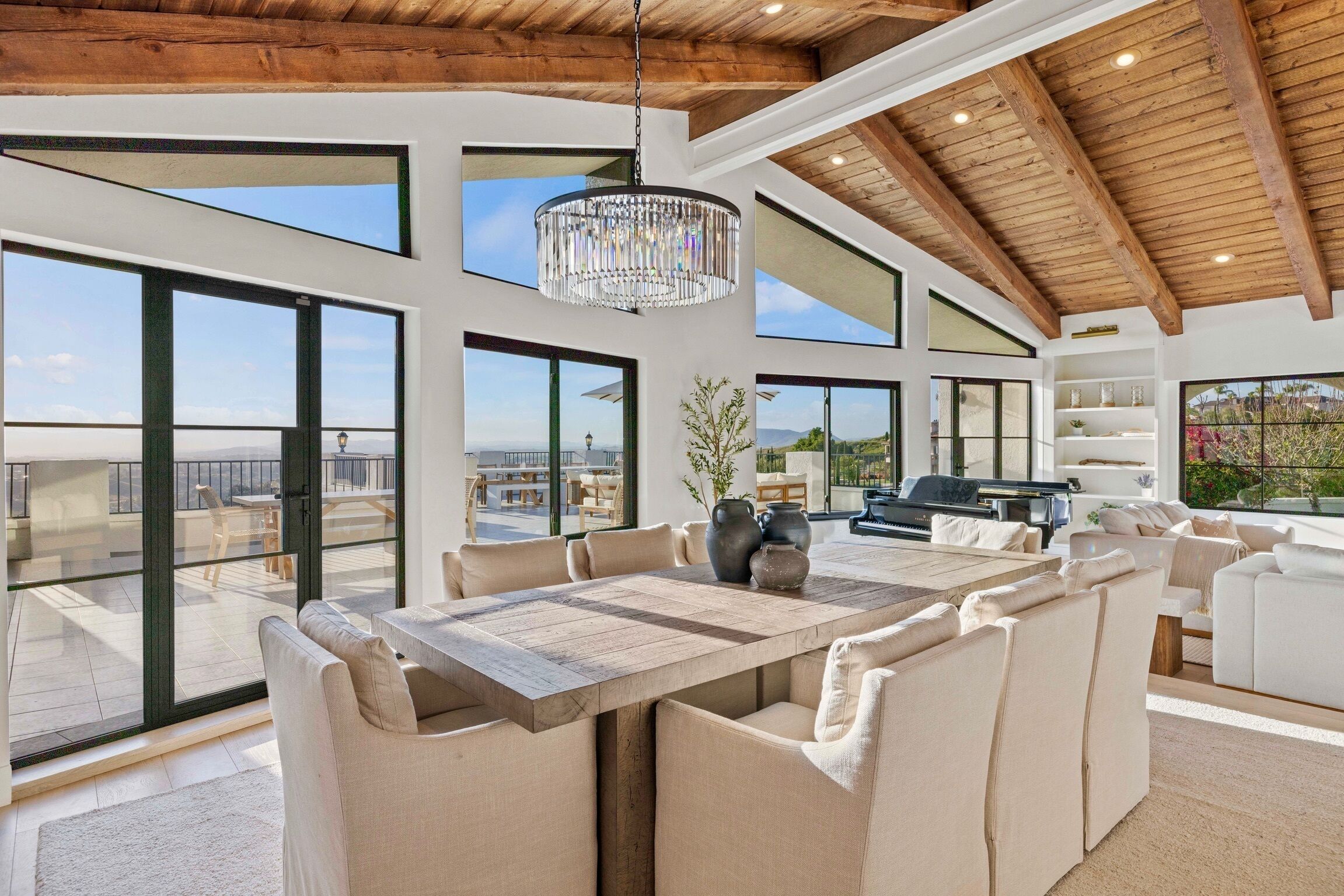 A spacious living area with a large wooden dining table surrounded by beige sofas, a chandelier, and large windows offering views of the outdoors.