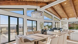 A spacious living area with a large wooden dining table surrounded by beige sofas, a chandelier, and large windows offering views of the outdoors.