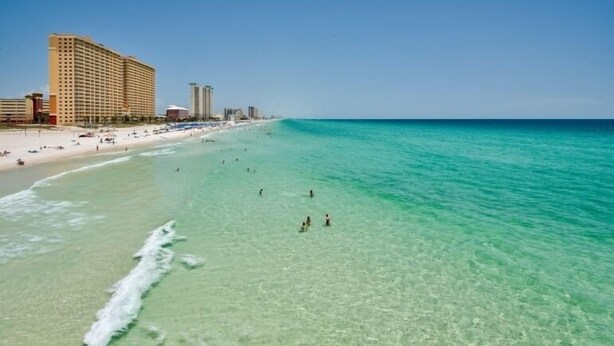 Una playa con agua turquesa cristalina, una orilla arenosa y varios edificios altos al fondo.