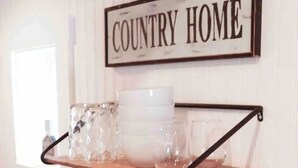 A kitchen with a 'COUNTRY HOME' sign, a wooden shelf with dishes and glasses, and a wicker basket with towels. - MaineStay Cottage #2 Bangor/Hampden close to town (Hampden)