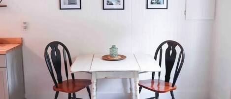 Un comedor con una mesa blanca y dos sillas negras con asientos de madera. Encima de la mesa, tres cuadros enmarcados en la pared.
