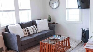 A living room with a grey sofa, a glass coffee table, a clock on the wall, and a television mounted above. - MaineStay Cottage #2 Bangor/Hampden close to town (Hampden)
