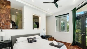 Une chambre moderne avec un grand lit, un ventilateur de plafond, un mur d’accent en bois, une vue à travers une porte vitrée et un escalier menant à un niveau supérieur.