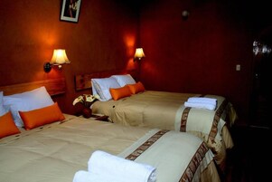 A hotel room with two beds, each with a beige bedspread and orange pillows. There are wall-mounted lamps on either side of the beds.