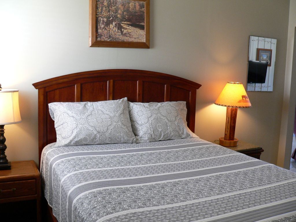 A neatly made bed with a patterned bedspread and pillows, a wooden headboard, a bedside lamp, and a framed picture on the wall.
