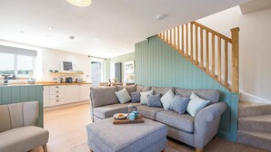 A modern living room with a grey sofa, a wooden staircase, and a kitchen area with a window. - Bwthyn Clyd, Holiday Cottage Llanrhian, Sleeps 4, 2 bedrooms,  bathrooms (Haverfordwest)