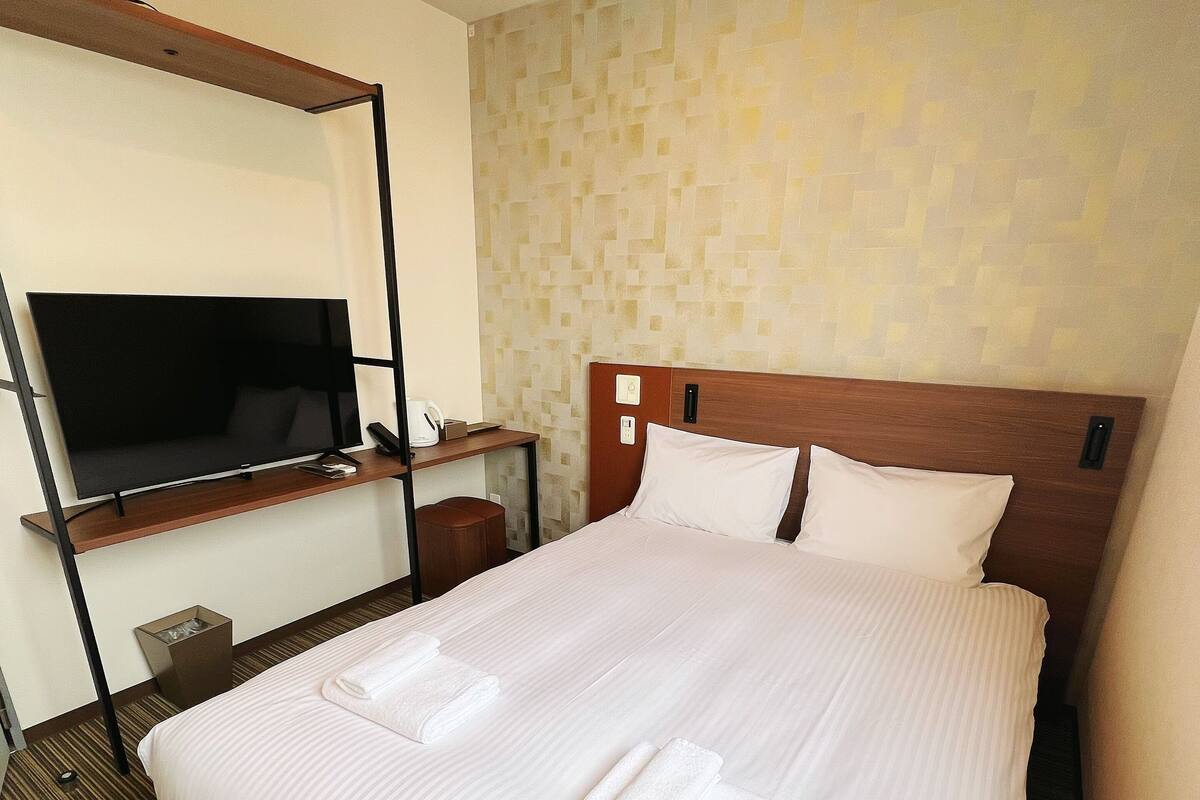 Habitación de hotel con una cama grande, un televisor de pantalla plana sobre un soporte de madera, un escritorio pequeño con una silla y un papel tapiz estampado.