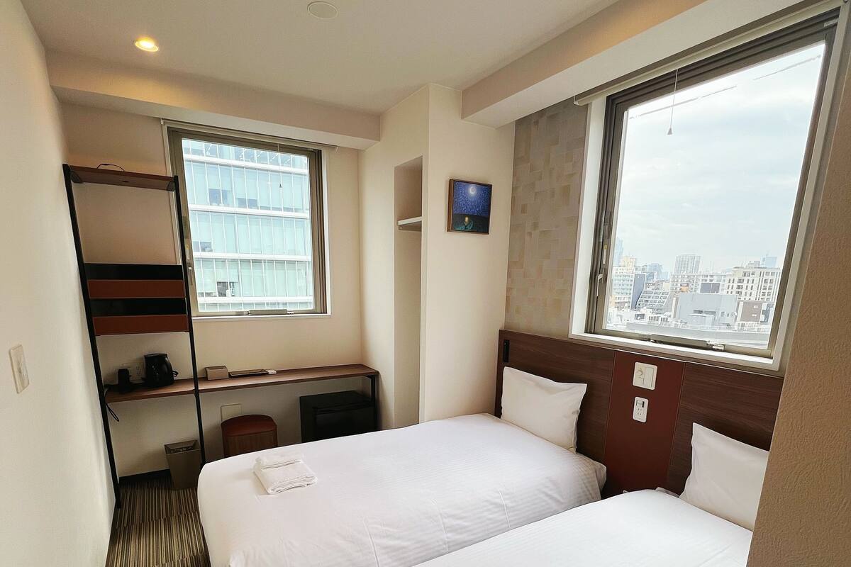 Habitación de hotel con dos camas, un escritorio, una silla, un ventanal con vista a la ciudad e iluminación empotrada en el techo.
