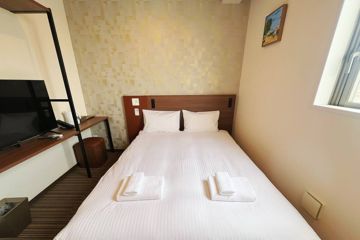 Una cama bien hecha con sábanas blancas y dos toallas dobladas, un televisor sobre un soporte de madera, y una pared con diseño de fondo.