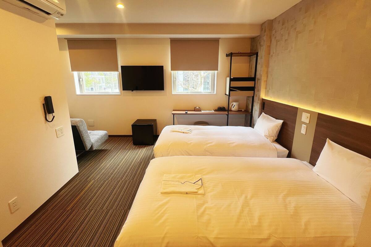 Una habitación de hotel con dos camas, un escritorio con silla, televisión y una ventana con persianas.
