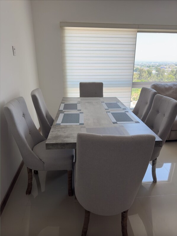 A dining area with a rectangular table and four chairs, a large window with blinds, and a view of the outdoors. - Departamento de lujo con alberca panoramica en Zona Metropolitana de Guadalajara (San Pedro Tlaquepaque)