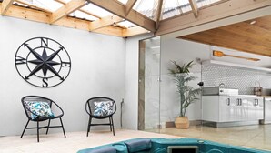 Un espace piscine intérieur moderne, doté d’un jacuzzi, de deux fauteuils avec des coussins aux motifs tropicaux, d’une grande décoration murale représentant une boussole et d’une cuisine visible à travers une porte en verre.