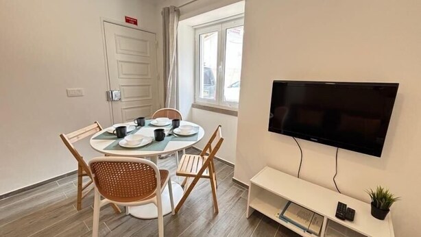 Una habitación pequeña y bien iluminada con una mesa de comedor preparada para cuatro personas, un televisor de pantalla plana en la pared y una pequeña consola blanca con una planta.