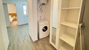 Habitación de hotel con lavadora, una pequeña estantería y una puerta que da a otra habitación.