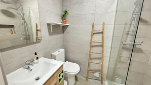 Baño con lavamanos, inodoro y ducha. Se ve una escalera apoyada en la pared y un estante pequeño con una planta.