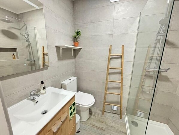 Une salle de bain équipée d’un lavabo, de toilettes et d’une douche. Une échelle est appuyée contre le mur, et une petite étagère porte une plante.
